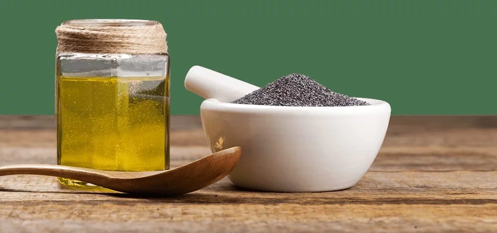 Bouteille en verre d’huile de nigelle dorée à côté d’un bol blanc avec des graines noires et une cuillère en bois.